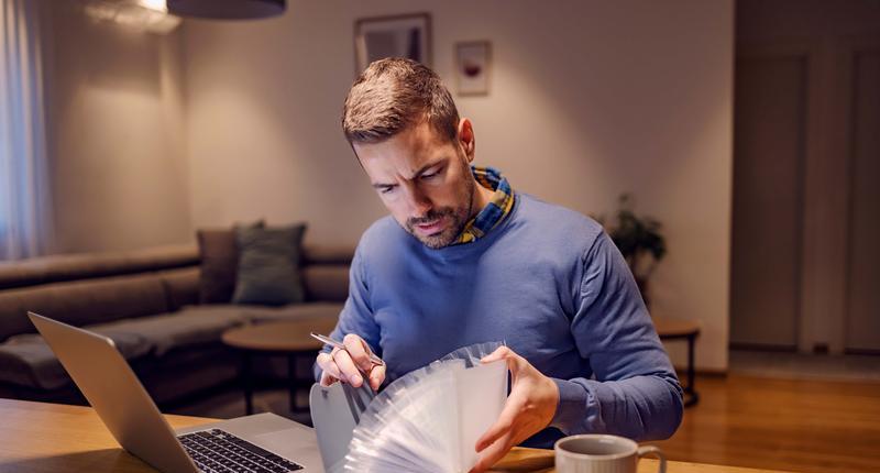 Finances bills family budget and economy. A young man sitting at home with a poly envelope in his hands and looking at bills. He is going to pay bills online on the laptop